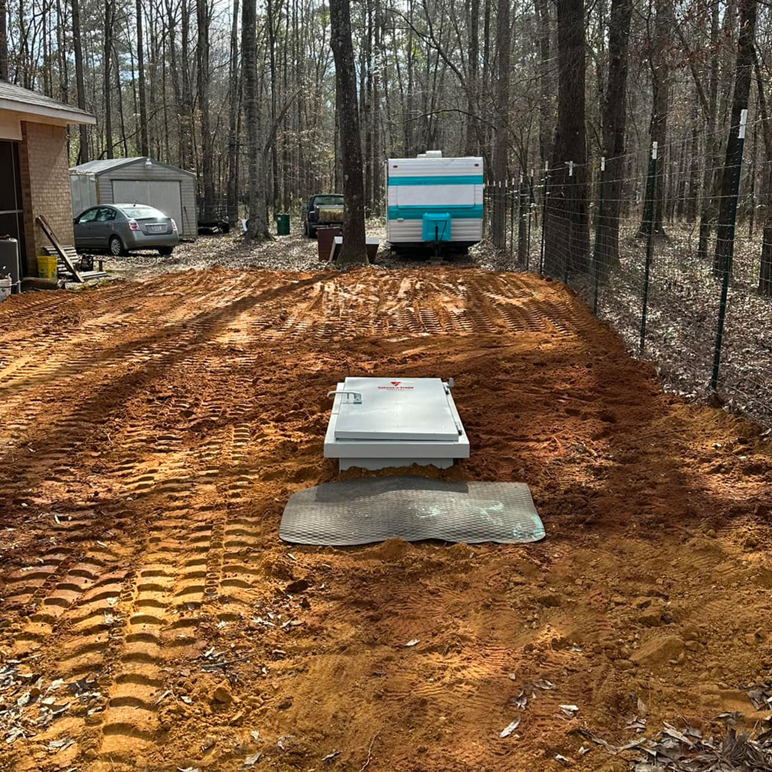Estate 10x6 Max - Storm Shelters of Iowa Backyard construction scene with orange dirt, a rectangular metal access hatch on the ground, and a trailer in the background.
