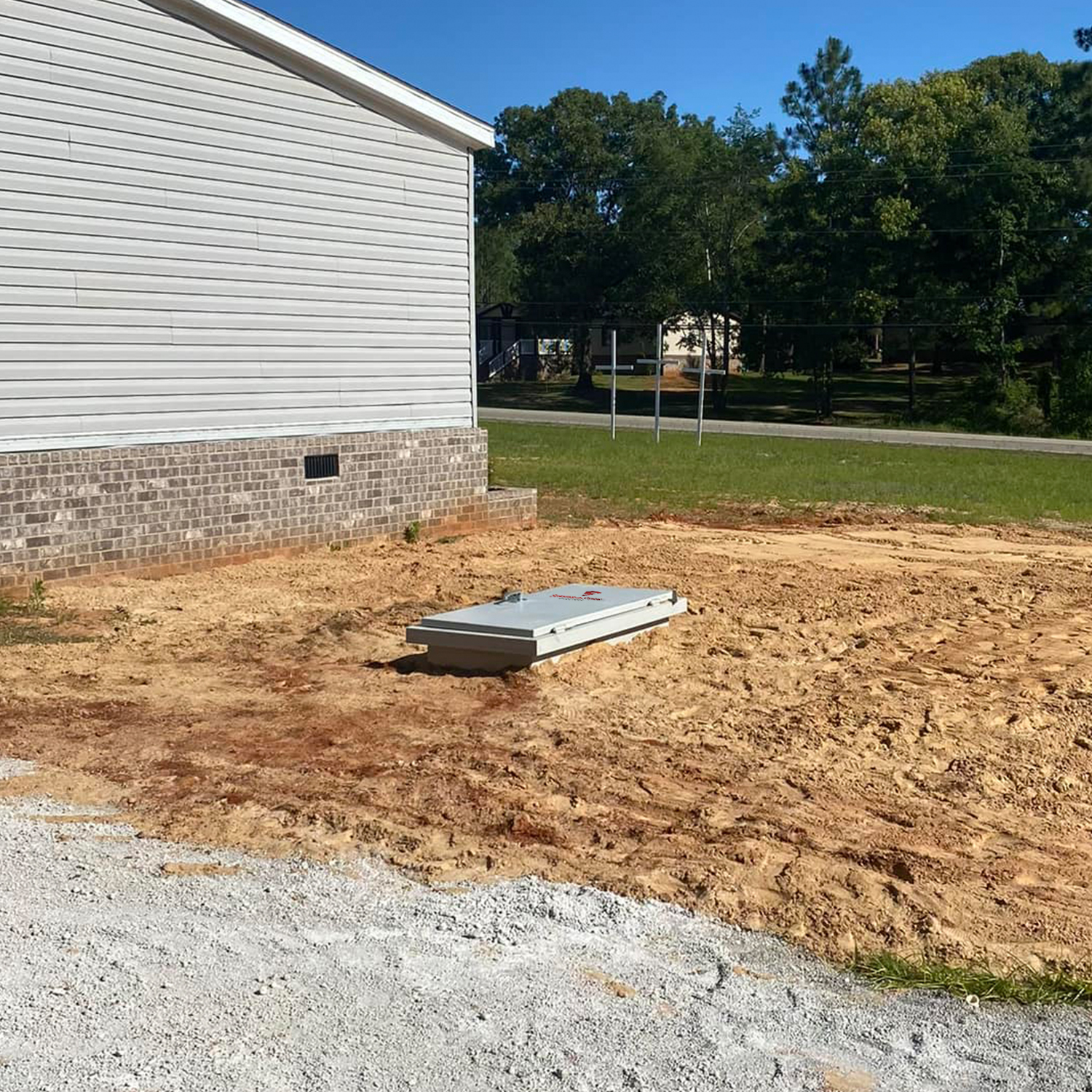 Estate 5x6 - Storm Shelters of Iowa Rectangular metal utility access hatch resting on dirt beside a light-silver house with brick foundation, in a clearing area.