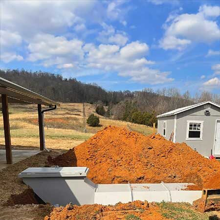 Construction site with a mound of orange dirt and concrete formwork in the foreground, a gray shed to the right, and rolling hills in the background.