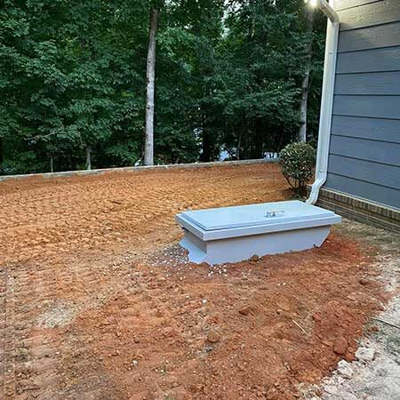 Gray rectangular utility access lid on exposed reddish dirt beside a house with a downspout, trees in the background
