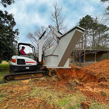 Compact excavator with an operator loading soil into a large metal dump hopper at a construction site.