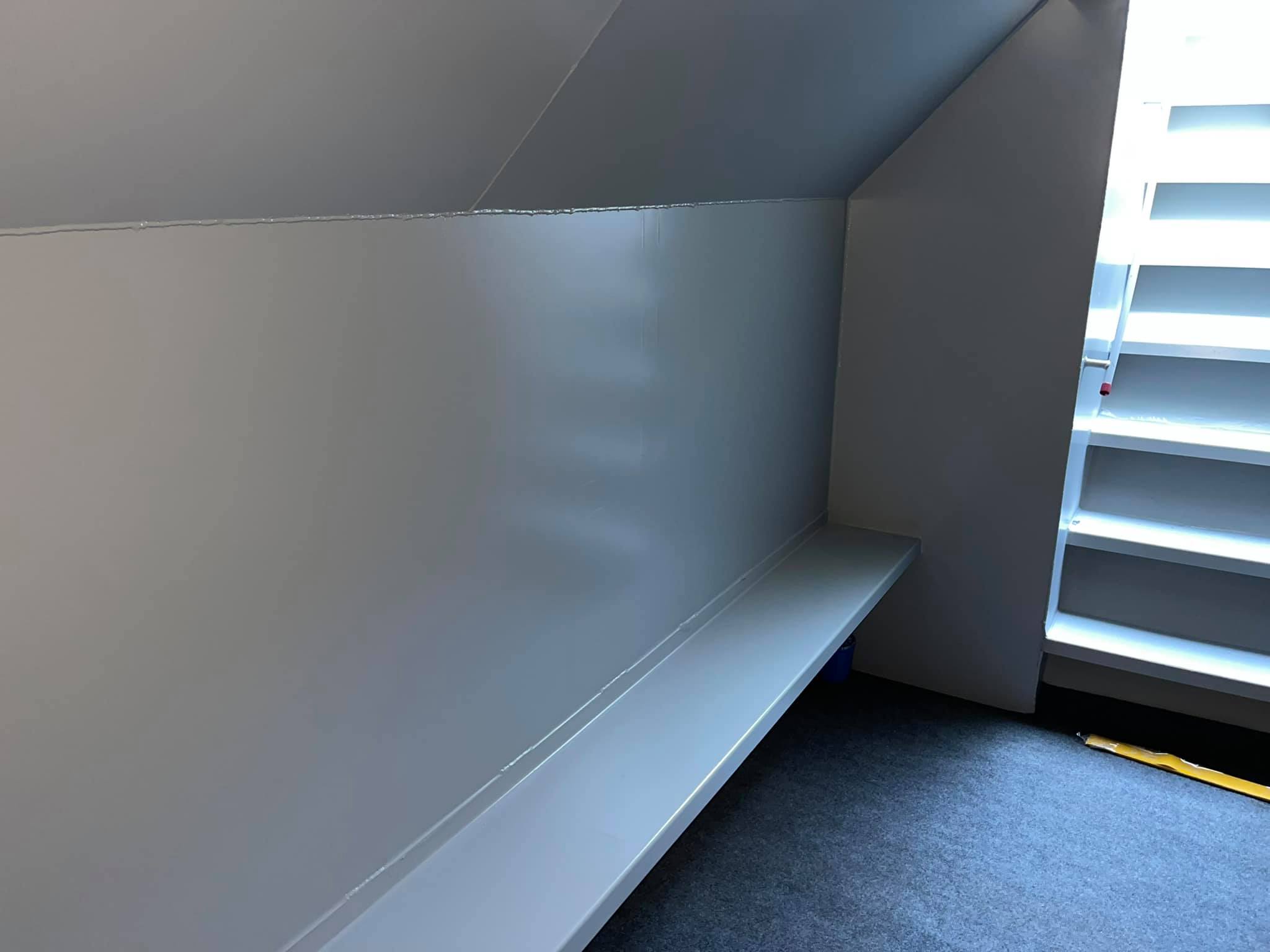 Corner of a gray storage room with a long white shelf along the wall and a slanted ceiling; bright white louvered window on the right side.
