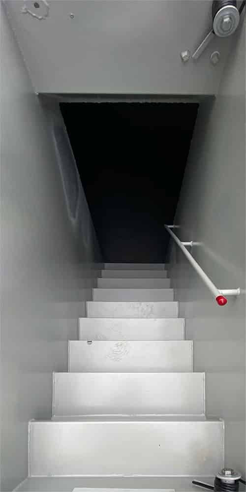 Narrow metal staircase descending into a dark basement, with a white handrail on the right side.