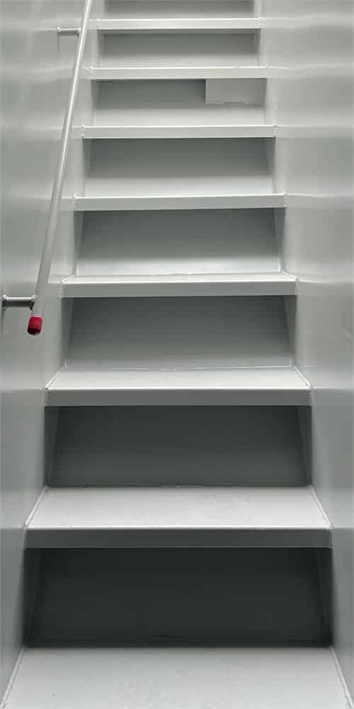 Indoor metal stairwell with light gray steps and a white handrail along the left; pink end cap on the railing is visible as the stairs rise away from the viewer.