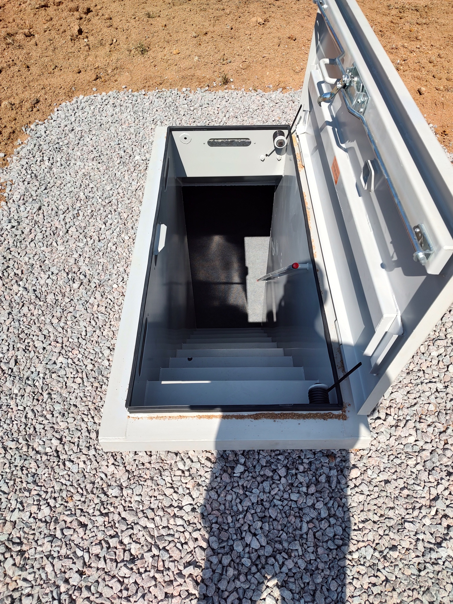 Estate Door - Storm Shelters of Iowa Open metal hatch with descending stairs into a dark underground area, set on gravel and dirt ground outside.”,